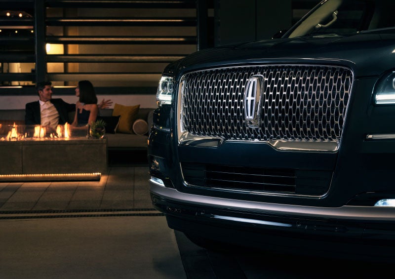 A close-up of a blue 2024 Lincoln Navigator grille and emblem, parked near a couple conversing by a firepit at night, seated on a bench outside a restaurant near Elizabethtown, Kentucky.