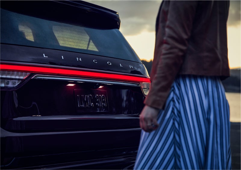A person is shown near the rear of a 2023 Lincoln Aviator® SUV as the Lincoln Embrace illuminates the rear lights | Don Franklin Lincoln Elizabethtown in Elizabethtown KY